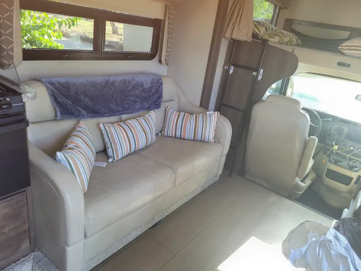 RV rental interior: a comfy couch with striped pillows, an overhead bunk bed, and the driver's cockpit area.. Jayco Greyhawk 2018