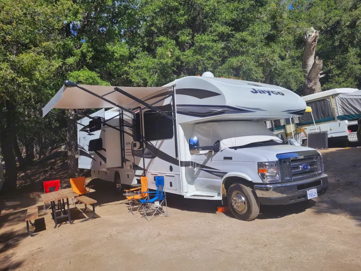 Class C RV rental at a sunny campsite with an extended awning, picnic table, and chairs among trees.. Jayco Greyhawk 2018