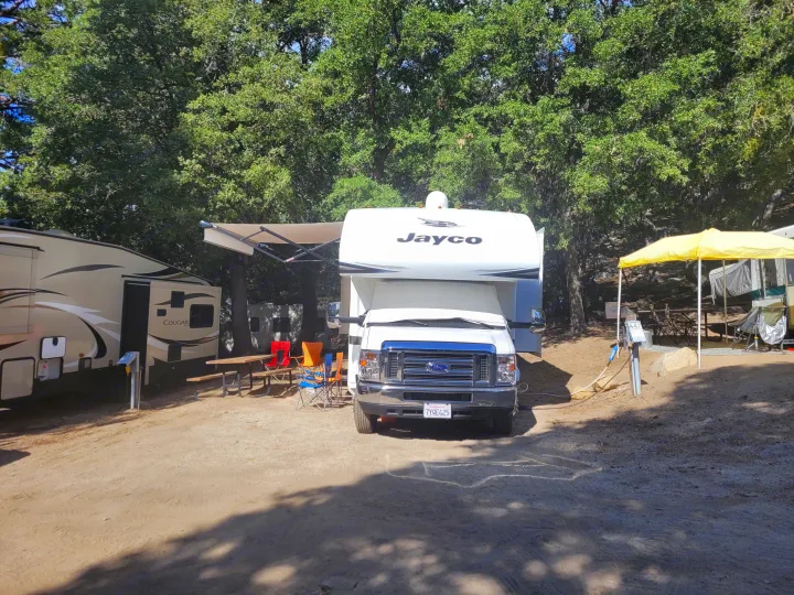 Relaxing RV rental site featuring a Jayco motorhome, travel trailer, picnic table, and yellow canopy amidst lush trees.. Jayco Greyhawk 2018