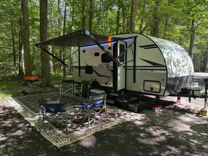 A modern rv rental travel trailer, with awning extended, set up at a shaded campsite with outdoor rug, grill, and chairs.. Venture Sonic Lite 2021