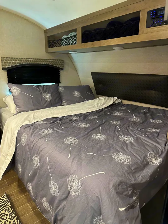Cozy RV rental bedroom features a bed with dandelion duvet, headboard, storage, and control panel for a comfortable stay.. Venture Sonic Lite 2021