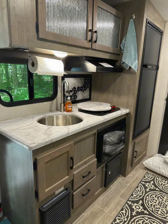 Inside a compact RV rental kitchen: sink, cooktop, microwave, fridge, cabinets, and a window overlooking trees.. Venture Sonic Lite 2021