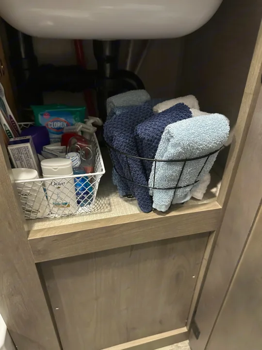 RV rental bathroom cabinet showcasing organized essentials: toiletries in a white basket and various blue & white towels.. Venture Sonic Lite 2021