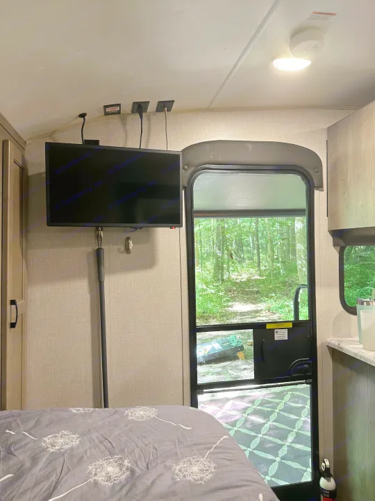 Inside a cozy RV rental bedroom featuring a mounted TV, a dandelion comforter, and a serene forest view from the door.. Venture Sonic Lite 2021
