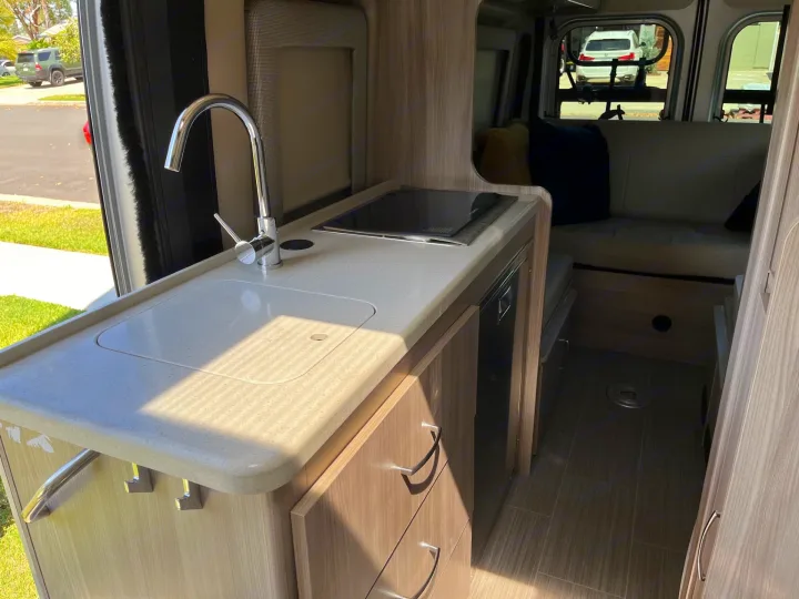 Inside a spacious RV rental: full kitchen with sink/cooktop, wood cabinets, and rear bench seating with pillows.. Thor Sequnce 2023