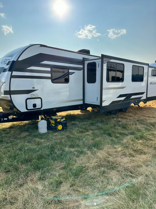 A white RV rental travel trailer with extended slide-out and generator, parked on a grassy field on a sunny day.. Heartland RVs Mallard 2022