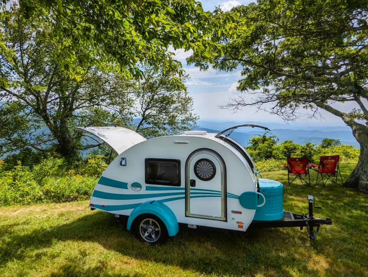 Enjoy mountain views with this white and teal teardrop camper RV rental, parked on grass near two red camping chairs under shady trees.. nuCamp T@G 2017