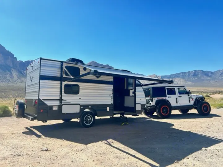 A white travel trailer (RV rental) with its awning out, parked next to a white Jeep in a desert mountain landscape.. Riverside Retro 190BH 2023