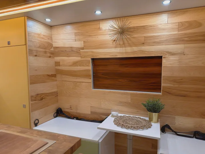 This RV rental interior showcases warm wood paneled walls, bright lighting, yellow cabinets, and compact seating with a table and plant.. Mercedes-Benz Sprinter 2022