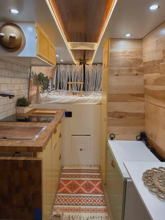 Cozy rv rental interior: compact kitchen, elevated bed with macrame, warm wood walls & cabinets. Functional and inviting.. Mercedes-Benz Sprinter 2022