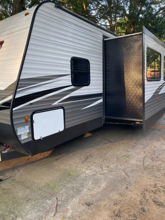 An rv rental travel trailer parked with its slide-out extended, showing a dark panel. Gray and white exterior.. Coleman Lantern 2021