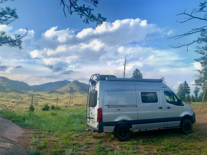 Silver Sprinter camper van, ideal for an rv rental, parked in a grassy mountain landscape under a bright, cloudy sky.. Winnebago Revel 2021