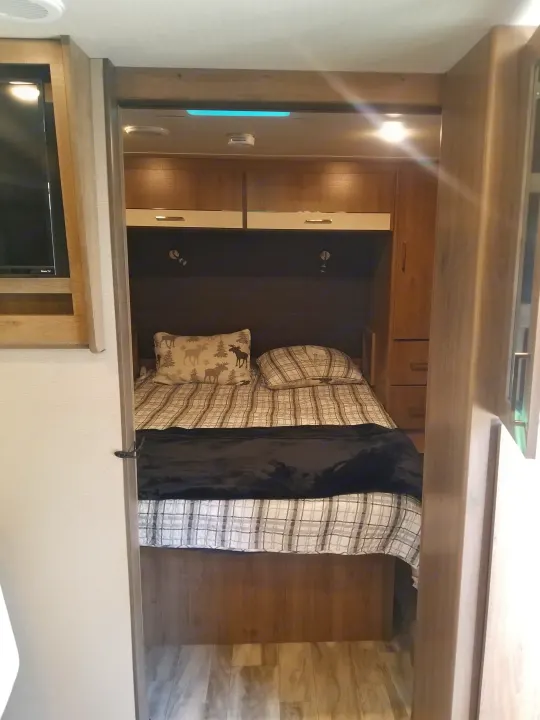 RV rental's cozy bedroom interior, featuring a plaid bed with moose pillow, overhead cabinets, reading lights, and side storage.. Grand Design Imagine XLS 2021