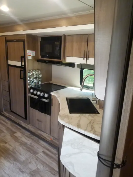 An RV rental kitchen interior showcasing a refrigerator, microwave, stove, sink, and light wood cabinetry with marble-look counters.. Grand Design Imagine XLS 2021