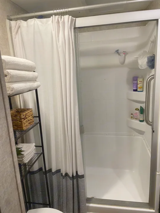 Clean RV rental bathroom featuring a shower with glass doors, white curtain, and a storage shelf with towels and toiletries.. Highland Open Range 2017