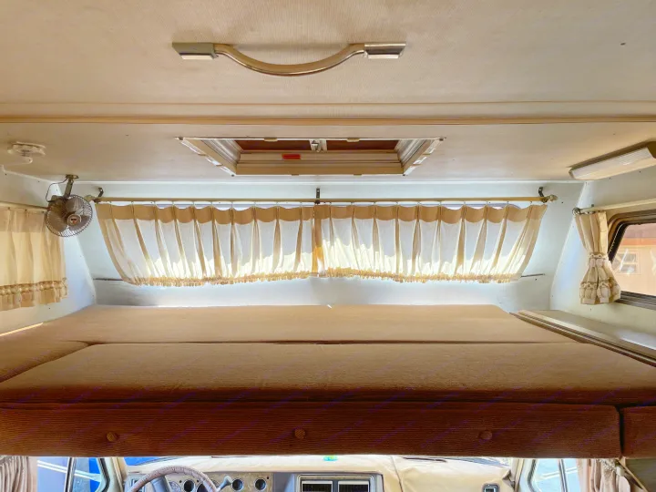 Looking into the cozy cab-over bunk area of an RV rental, showing curtains, a fan, and a roof vent.. Chevrolet Lazy Daze 1985