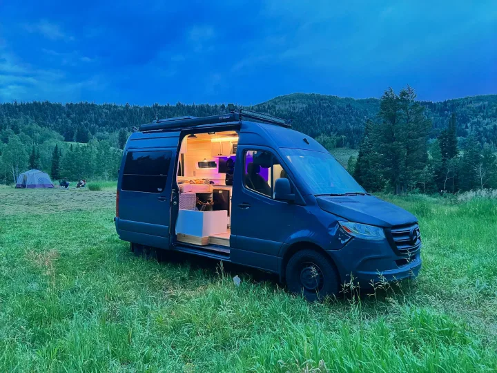 Twilight camping scene: A dark Mercedes Sprinter RV rental camper van with open, lit interior, tent, and mountains visible.. Mercedes-Benz Sprinter RV Motorhome Campervan 2019