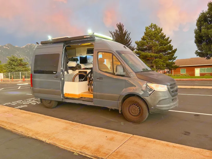 Custom gray camper van, an RV rental, with open side door revealing interior & bike. Parked outdoors under a sunset sky with mountains.. Mercedes-Benz Sprinter RV Motorhome Campervan 2019