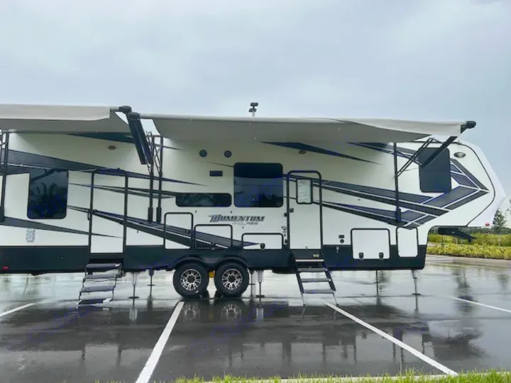 This white Momentum G-Class fifth-wheel RV rental is parked on a wet lot with awnings deployed, ready for your next trip.. Grand Design Momentum 2018