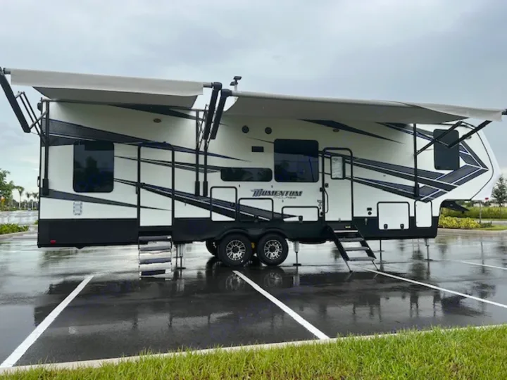 White fifth-wheel camper with extended awnings and steps down, parked on wet asphalt. Consider this for rv rental.. Grand Design Momentum 2018