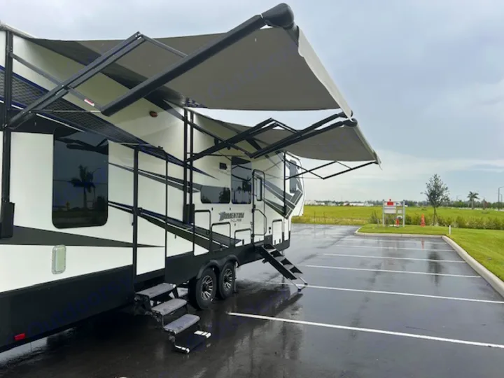 Modern RV rental, white and black with multiple awnings extended, parked in a wet lot on an overcast day.. Grand Design Momentum 2018