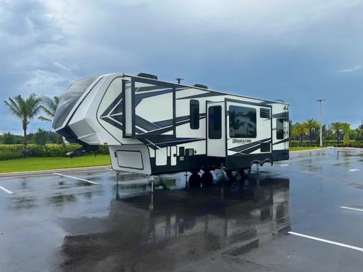 Modern white & black fifth-wheel RV, ideal for rv rental, parked on a wet lot with palm trees in the background.. Grand Design Momentum 2018