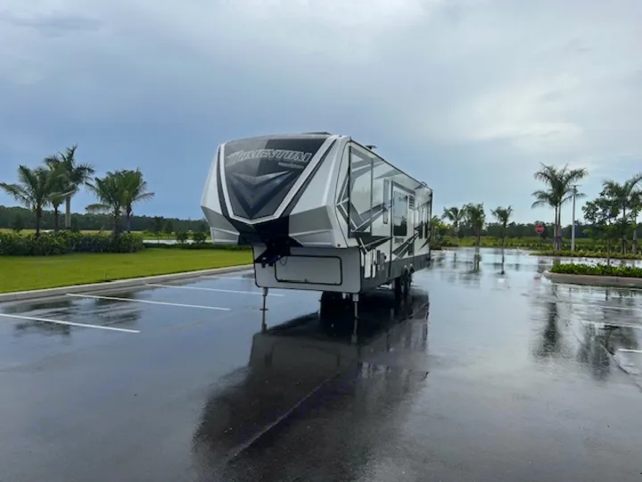 This sleek fifth-wheel RV rental is parked on a wet lot after rain, surrounded by lush palm trees. Ready for travel!. Grand Design Momentum 2018