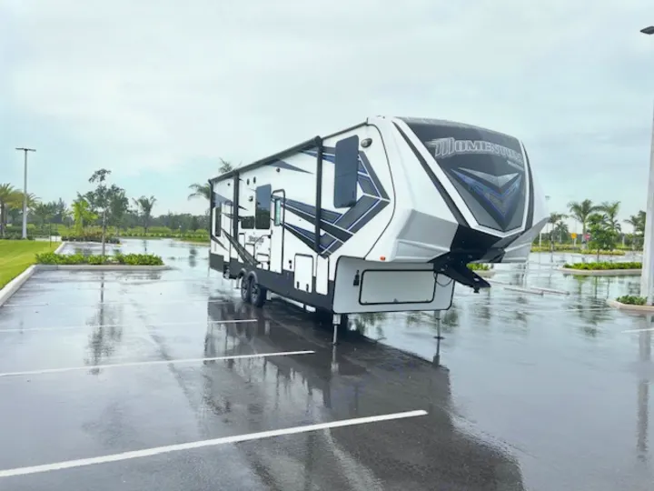 A white and black Momentum fifth-wheel RV, ready for rv rental, is parked on a wet asphalt lot, reflecting the overcast sky.. Grand Design Momentum 2018