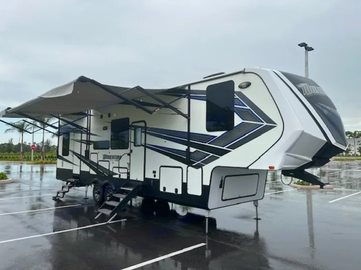 Large white and black Momentum fifth-wheel RV with awning extended, ready for rv rental. Parked in a wet lot.. Grand Design Momentum 2018