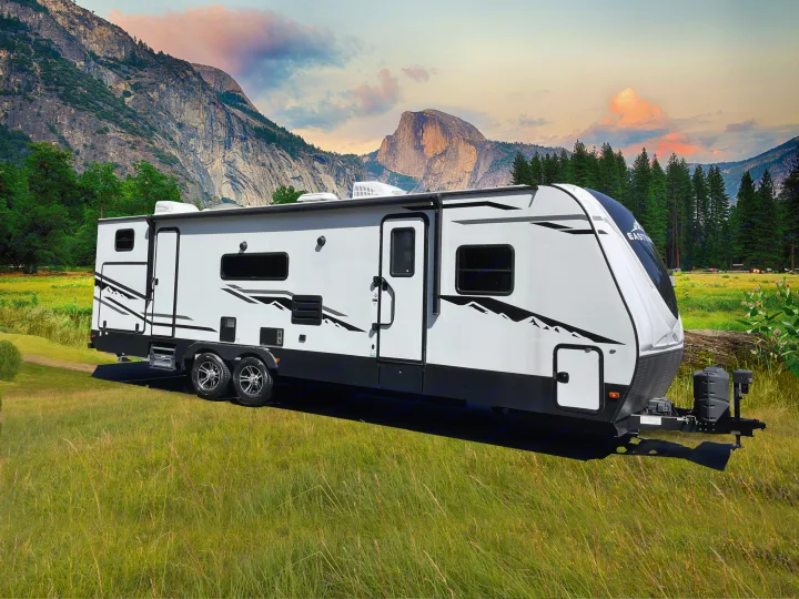 This white travel trailer **rv rental** is parked in a green meadow with towering mountains and a colorful sky, ideal for outdoor adventures.. East to West Alta 2021