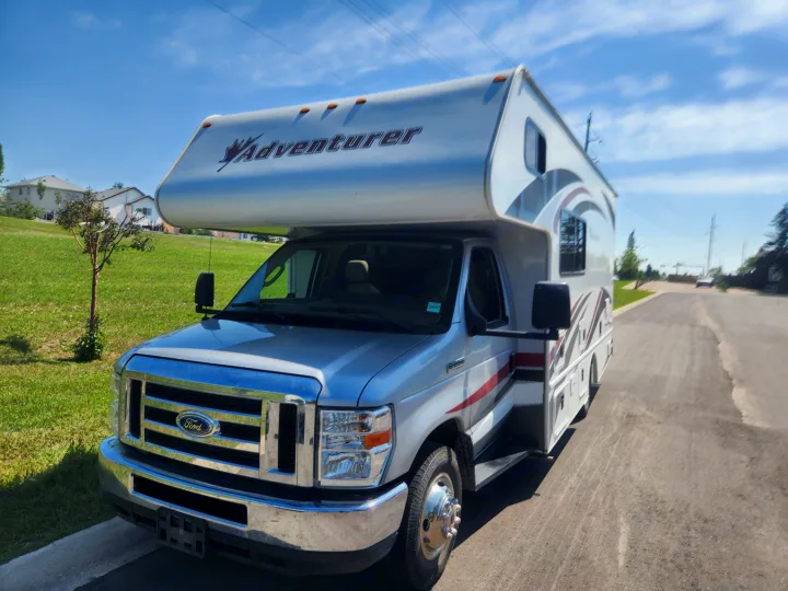 Front and side view of a silver Ford Class C Adventurer RV rental parked on an asphalt road beside a grassy hill.. Adventurer Adventurer Motorhome 2017