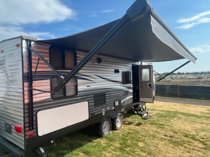 Travel trailer RV rental with a large dark awning extended, parked on green grass. Ideal for camping trips.. Keystone RV Springdale 2019