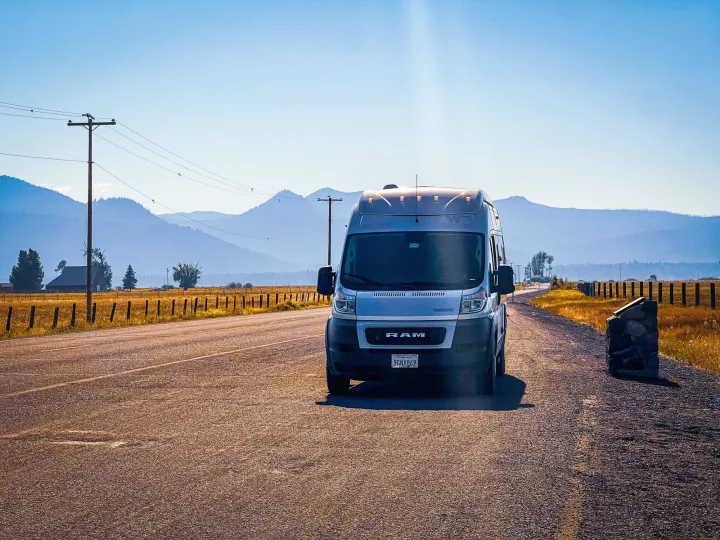 A white camper van (RV rental) rests on a sunlit rural road, with golden fields, fences, and blue mountains in the distance. Ideal for scenic road trips.. Winnebago Solis 2021