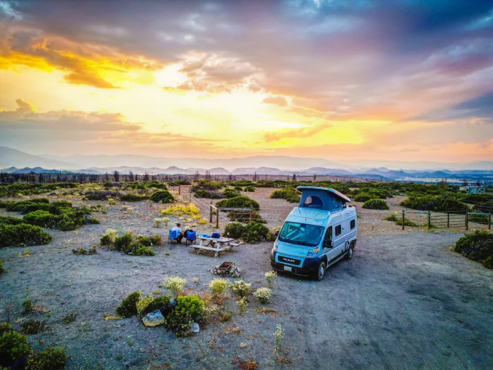 Scenic RV rental camping at sunset. A camper van, picnic table, and people are set up in a vast landscape.. Winnebago Solis 2021