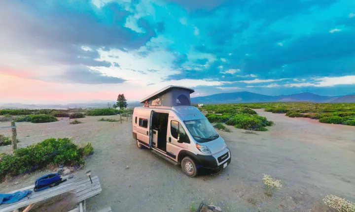 Enjoy an RV rental adventure! Silver camper van with pop-top at dusk, parked by a picnic table amidst mountains & colorful sky.. Winnebago Solis 2021