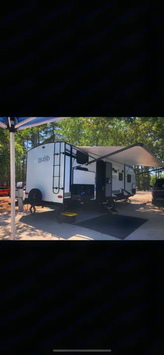 White Legend travel trailer RV rental, set up at a shaded campsite with an extended awning and outdoor mat.. Forest River Legend 2019