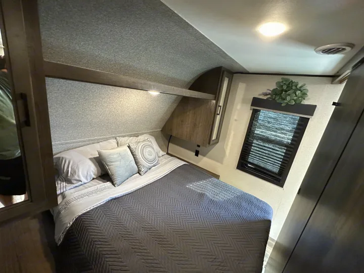 Inside an RV rental, a compact bedroom with a neatly made bed, gray linens, overhead storage, and a window with blinds.. Starcraft Autumn Ridge 2022