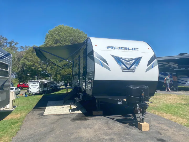 This white and blue Rogue by Forest River travel trailer, an RV rental, is set up at a sunny campsite with its awning extended.. Forest River Vengeance 2021