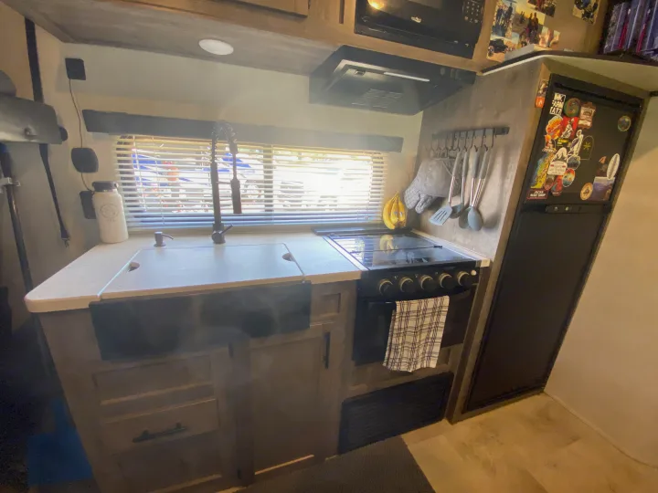 Compact RV rental kitchen with a sink, stove, microwave, refrigerator, hanging utensils, and a window.. Forest River Vengeance 2021