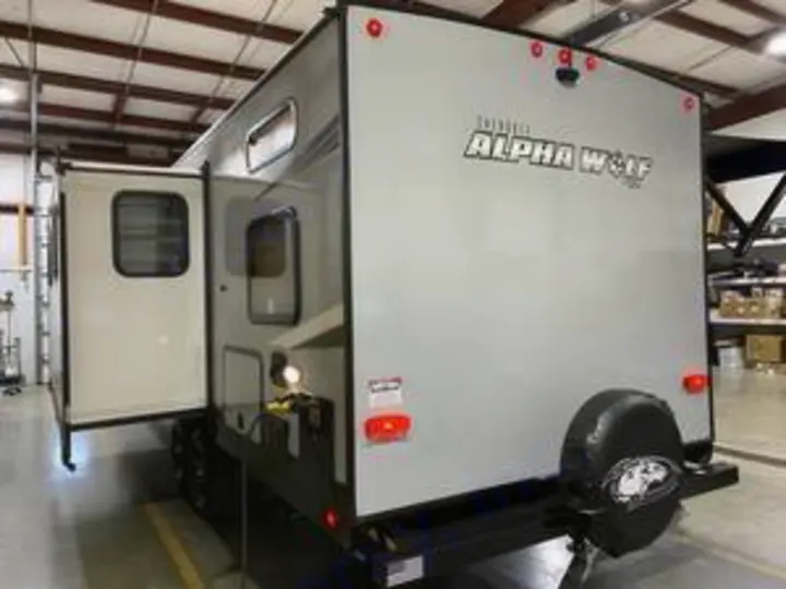Explore this Alpha Wolf travel trailer, a spacious RV rental. Its rear view shows a spare tire & open side door, inside a warehouse.. Forest River Cherokee Alpha Wolf 2022