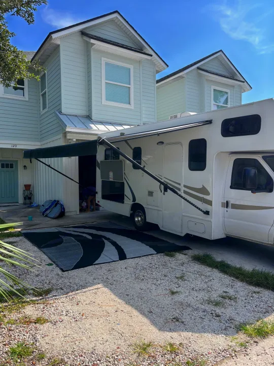 An RV rental is parked at a light blue house, its awning extended over an outdoor rug. Someone accesses an RV storage compartment.. Thor Majestic 2017