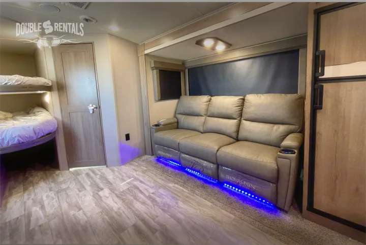Cozy rv rental interior featuring a plush reclining sofa with cool blue LED lights, bunk beds, and a refrigerator.. Grand Design Imagine 2021