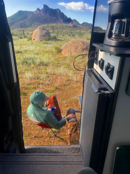 From an RV rental, a person in a hoodie and dog relax, facing mountains. Coffee maker inside. Scenic camping.. Winnebago Revel 2021