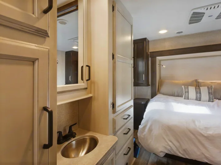 Inside a modern RV rental, showing a cozy bedroom with bed and an adjacent compact bathroom vanity with sink.. Entegra Coach Odyssey 2021
