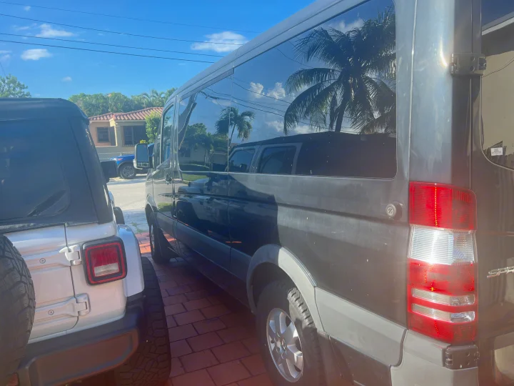 View of a dark RV rental Sprinter van with reflective windows showing palm trees, parked on brick pavers next to a white Jeep.. Mercedes Sprinter 2016