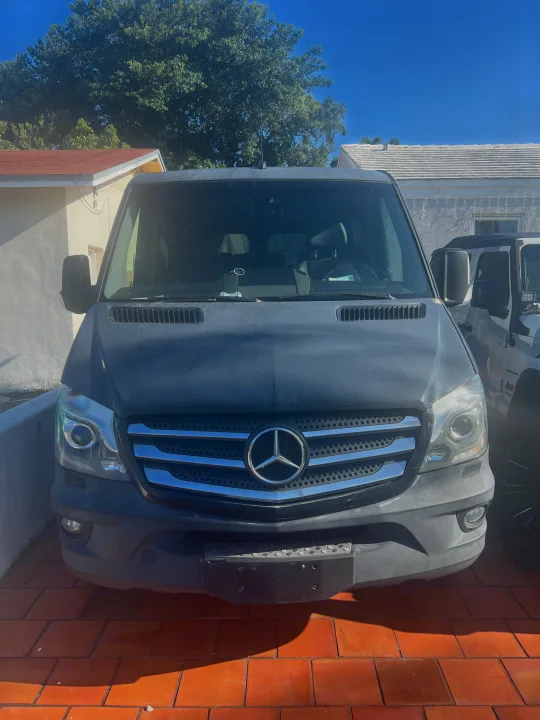 Front of a dark grey Mercedes-Benz Sprinter van, an RV rental, parked on reddish tiles. Blue sky, trees & houses visible.. Mercedes Sprinter 2016