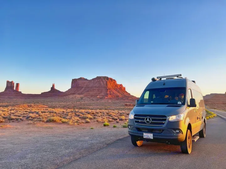 A gray Mercedes Sprinter RV rental van sits on a desert road, facing Monument Valley's red rock formations at sunset.. Thor Tranquility 2022