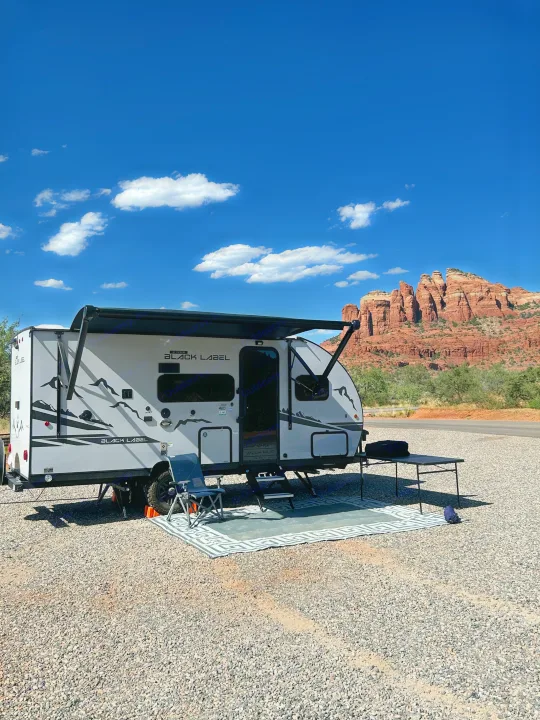 Stunning rv rental setup: a modern travel trailer with awning, outdoor rug, and chair at a gravel campsite with red rock mountains.. Forest River Wolf Pup 16 BHS Black Label 2023