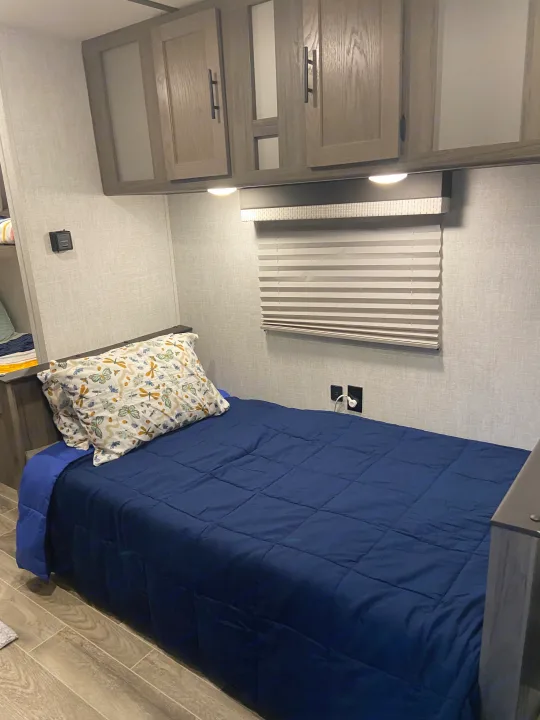 Cozy RV rental bunk bed with blue comforter, butterfly pillow, overhead cabinets, and window with blinds.. Gulf Stream Enlighten 2021