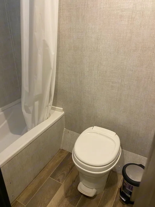 Compact RV rental bathroom with white shower/tub & curtain, foot-pedal toilet, and waste bin. Grey textured walls, wood-look floor.. Gulf Stream Enlighten 2021